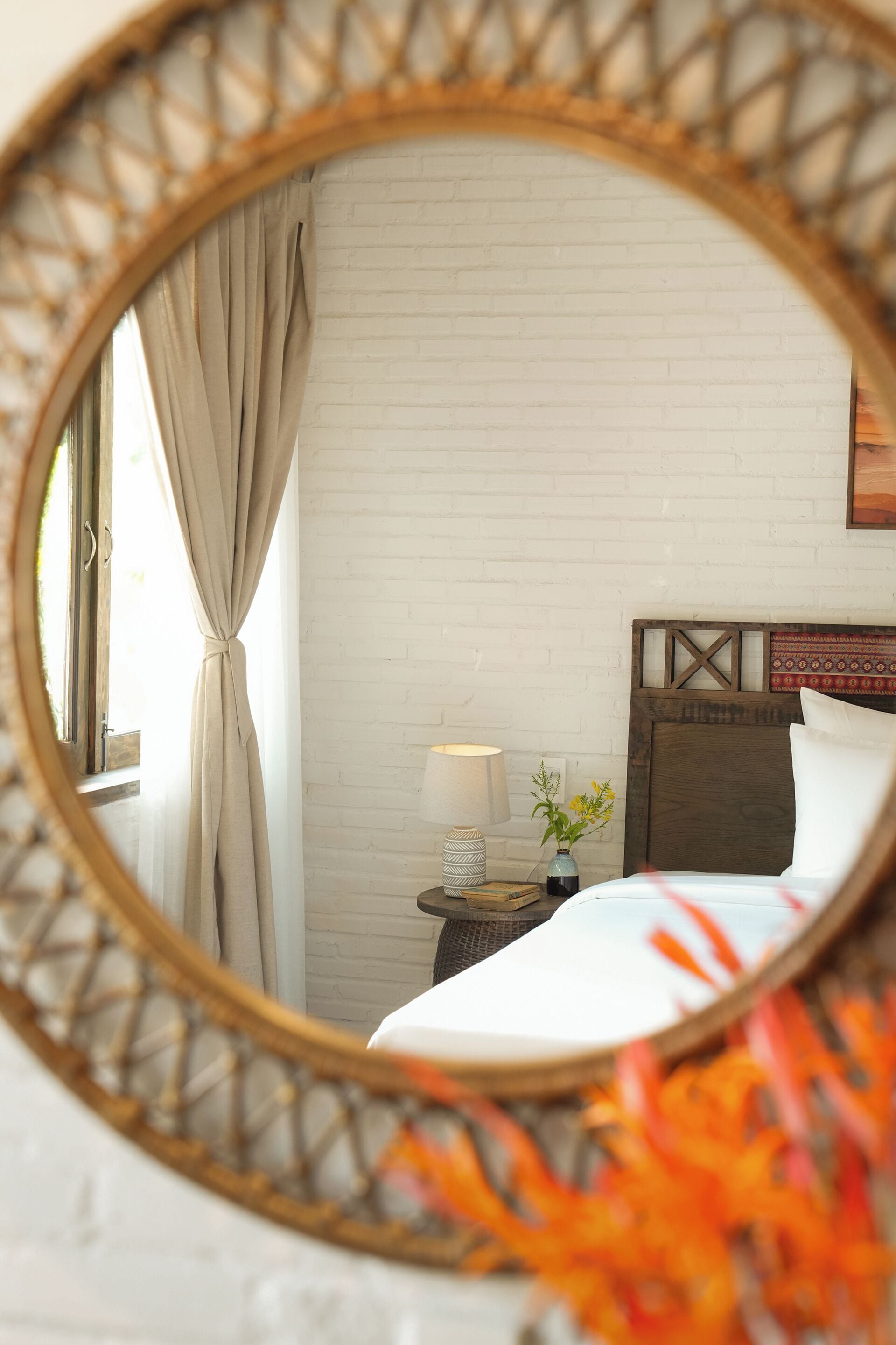 A bedroom with a white brick wall, a wooden headboard, a bedside table with a lamp, and a window with curtains.