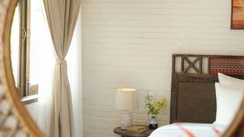 A bedroom with a white brick wall, a wooden headboard, a bedside table with a lamp, and a window with curtains.