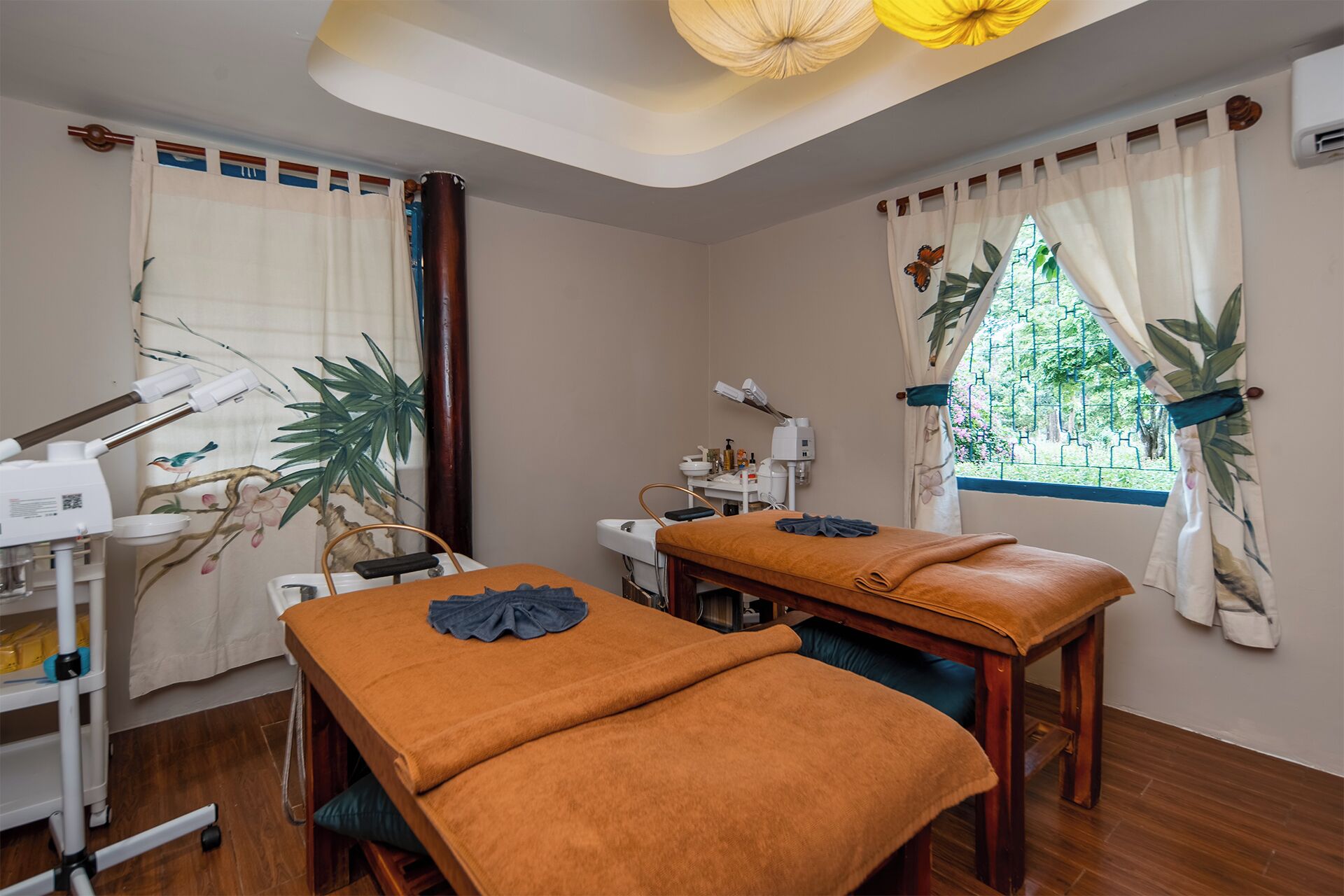A room with two massage tables, a ceiling light fixture, a window with curtains, and a wall-mounted air conditioning unit.