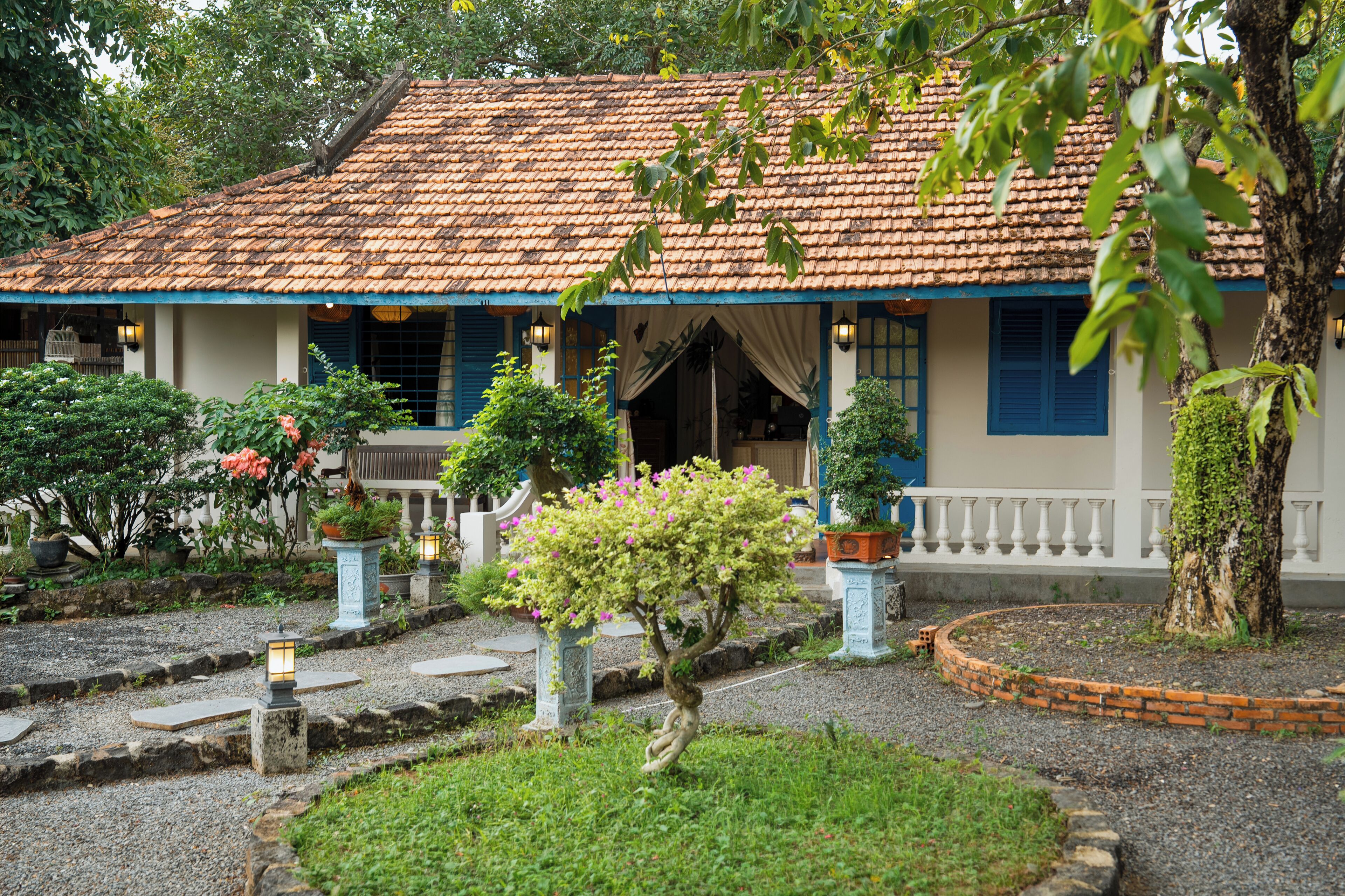 Una casa tradicional con techo de tejas, un porche cubierto y un jardín con plantas en macetas y un pequeño árbol.