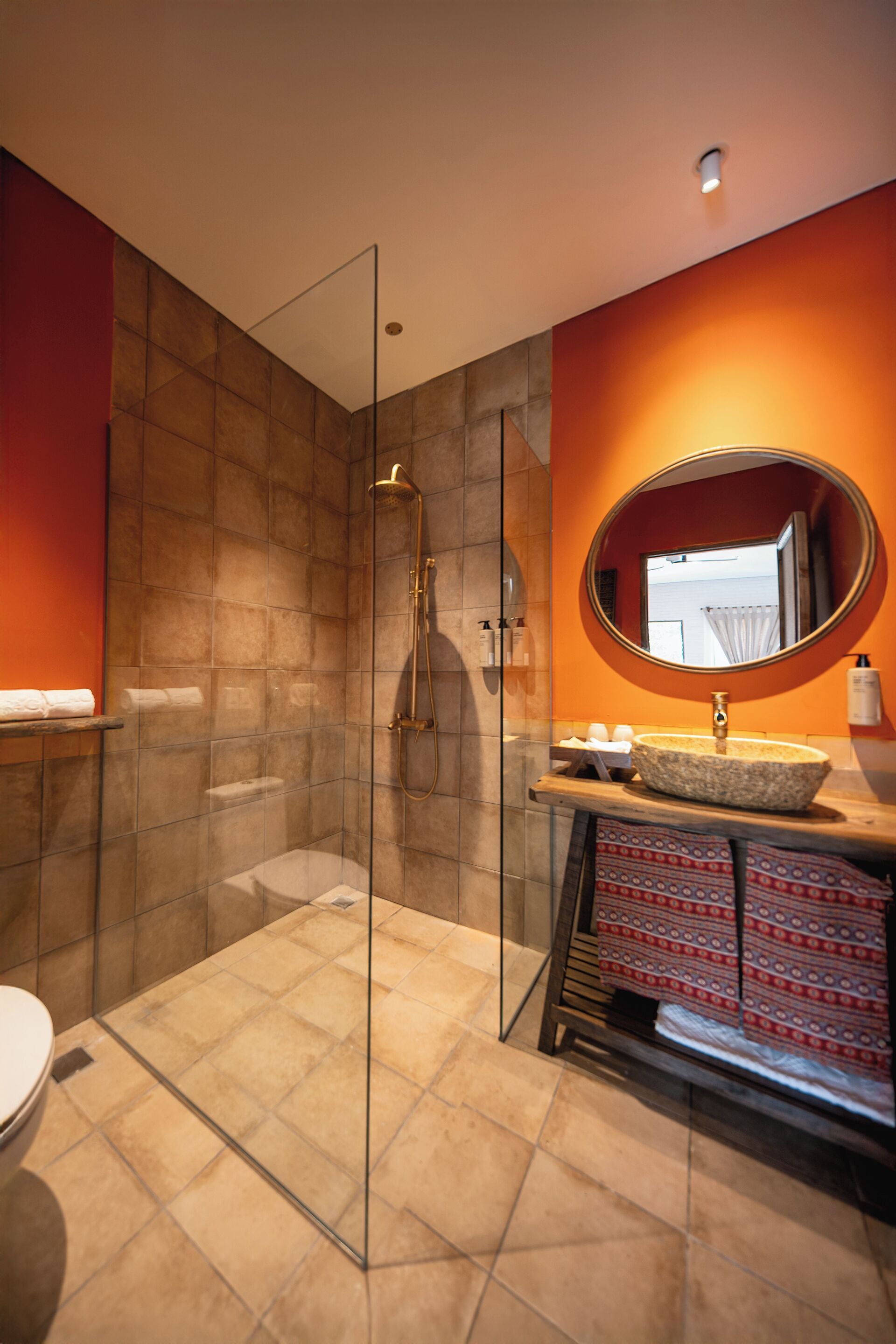 A modern bathroom with a glass shower enclosure, a round mirror, and a beige tiled floor.