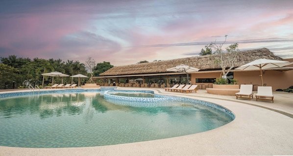 A swimming pool with lounge chairs and umbrellas, surrounded by a building with a thatched roof. - Alagon Ecolodge Nam Cat Tien (Nam Cat Tien)