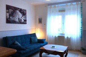Ein Wohnzimmer mit einem blauen Sofa, einem Holztisch, einem Bild an der Wand und einem Fenster mit Vorhängen.