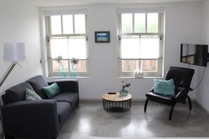 Ein Wohnzimmer mit einer Couch, einem Sessel, einem runden Couchtisch und zwei Fenstern mit Jalousien.