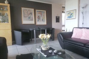 Una sala de estar con una mesa de centro de vidrio, un sofá negro, una mesa de comedor y obras de arte en las paredes.