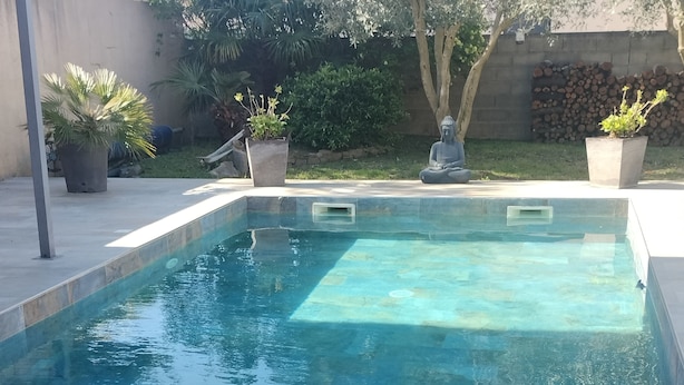 Et rektangulært swimmingpool med flisekant, omgivet af en have med krukkeplanter og en stenvæg.