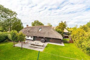 Ein reetgedecktes Haus mit Terrasse, umgeben von einem gepflegten Rasen und Bäumen.