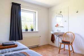 Ein Schlafzimmer mit Bett, einem Fenster mit Vorhängen, einem Heizkörper, einem Holzstuhl und einem Badezimmer mit Waschbecken und Spiegel.