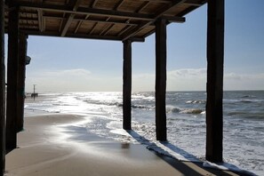A beach view through a wooden structure with columns, sandy beach, and ocean waves. - Holiday home with 2 bedrooms (Callantsoog)