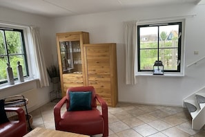 A room with a wooden cabinet, a red chair, a dining table, and a window with a view of greenery. - Holiday home with 2 bedrooms (Callantsoog)