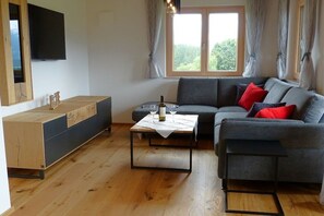 Ein Wohnzimmer mit einer Couch, einem Couchtisch, einem an der Wand montierten Fernseher und einem Fenster mit Vorhängen.