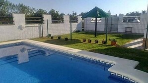 Una piscina con zona vallada, un pequeño jardín y un pabellón.
