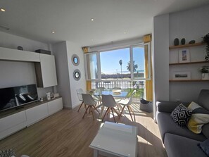 Ein modernes Wohnzimmer mit einem Flachbildfernseher, einem Glastisch und einer Couch mit gemusterten Kissen.