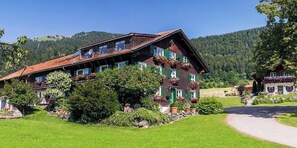 Ein traditionelles Chalet mit geneigtem Dach, umgeben von üppiger Vegetation und einer Bergkulisse.