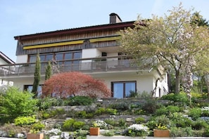 Ein mehrgeschossiges Gebäude mit Balkon, umgeben von einem angelegten Garten mit verschiedenen Pflanzen und einer Steinmauer.