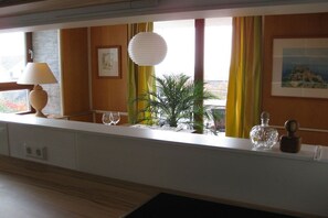 Ein Zimmer mit Fensterblick, einer Lampe, einem Topfpflanzen und einem gerahmten Bild an der Wand.