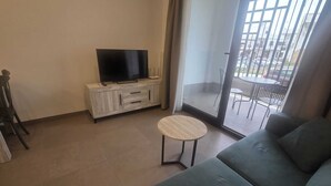 Un soggiorno con un divano, un tavolino rotondo, una televisione su un mobile e una porta vetrata scorrevole che dà su una zona esterna con sedie.