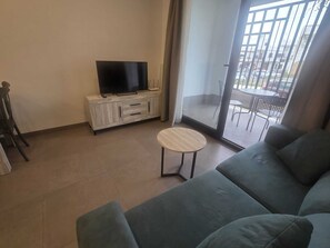 Ein Wohnzimmer mit einer Couch, einem kleinen runden Tisch, einem Fernseher auf einer Anrichte und einer Glastür, die zu einem Außenbereich mit Stühlen führt.