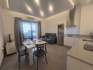 Ein kompakter Wohnraum mit Esstisch, Stühlen, einer Couch, einer Küchenzeile und Blick durch ein Fenster.