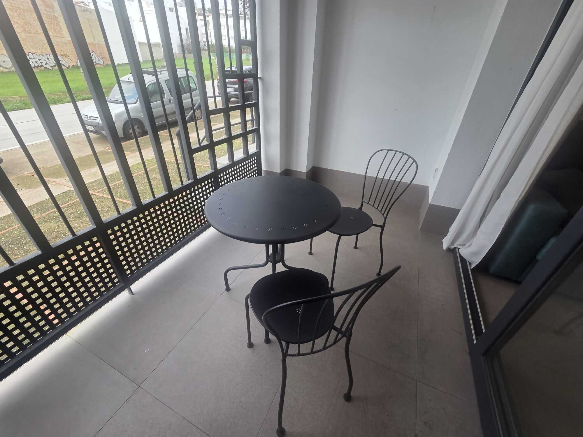 Ein Balkon mit rundem Tisch und zwei Stühlen, Blick auf geparkte Autos und eine Metallzäunung.