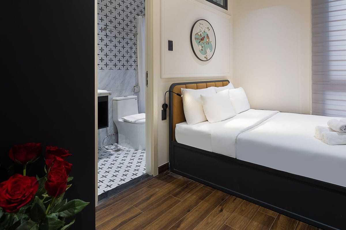 Une chambre à coucher comprenant un lit, une table de chevet, une fenêtre avec des stores, une salle de bain avec des toilettes et un mur orné d’une assiette décorative.