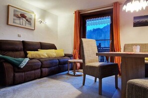 Ein Wohnzimmer mit einer braunen Ledersofa, einem kleinen runden Tisch, einem Korbstuhl und einem Fenster mit orangefarbenen Vorhängen.