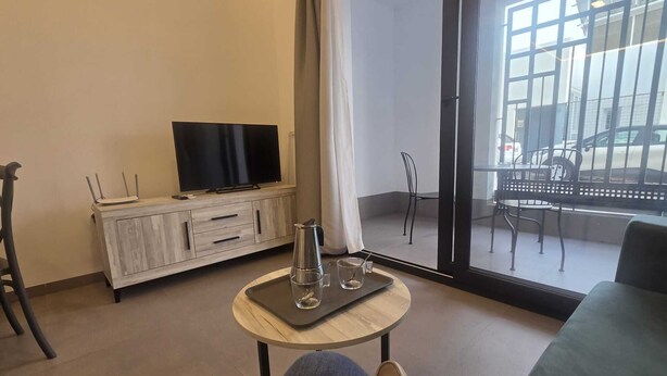 A modern living room with a television on a wooden cabinet, a small round table with a glass, and a view of the street through large glass doors. - Apartment 2 Virgen de la Luz (Conil De La Frontera)