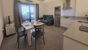 A compact living space with a dining table, chairs, a sofa, a kitchenette, and a view through a window. - Apartment 2 Virgen de la Luz (Conil De La Frontera)