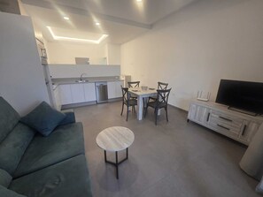 Ein modernes Wohnzimmer mit Sofa, Esstisch mit Stühlen, Fernseher und Küchenzeile inklusive Schränken und Geschirrspüler.