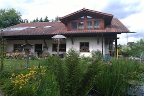 Ein Haus mit geneigtem Dach, mehreren Fenstern und einem Garten mit gelben Blumen und Grünpflanzen.