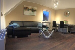 A living room with a black sofa, a glass coffee table, and a dining area with a table and chairs. - Holiday apartment with 2 bedrooms (Uhldingen-Mühlhofen)