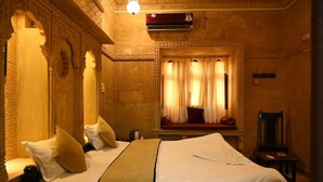 A hotel room with two beds, a window with curtains, a wall-mounted air conditioner, and a small table with a chair. - hotel kalyan haveli jaisalmer (Jaisalmer)