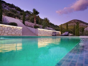 Piscina com degraus de pedra que levam a uma área de estar, cercada por vegetação e uma montanha ao fundo.