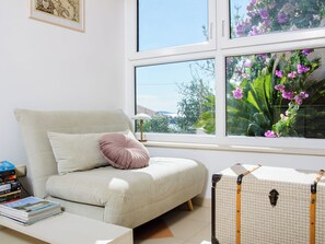A room with a sofa, a table with books, a suitcase, and a window with a view of greenery and flowers. - Vacation home Nerium by Interhome (Trogir/Vranjica)