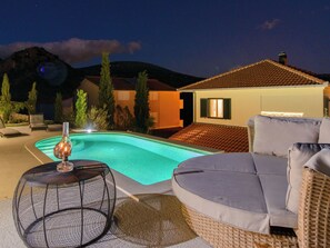 En swimmingpool med en solstol och ett runt bord, omgiven av ett hus med ett tak täckt av tegelpannor och ett berg i bakgrunden.