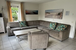 A living room with a sectional sofa, a coffee table, and a chair. There is a window with curtains, a painting on the wall, and a cabinet.