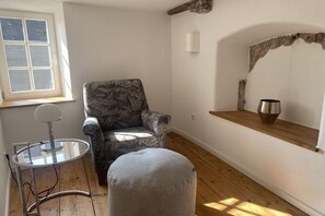 Ein Zimmer mit einem gemusterten Sessel, einem runden Hocker, einem kleinen Tisch mit Lampe, einer Holzablage und einem Fenster.