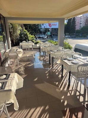 Un espacio exterior para comer con mesas y sillas, vista a la calle y un edificio con un cartel de fondo.