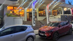 Een hotelentree met een trap die naar de lobby leidt, een bord met de naam van het hotel, en een rode auto die ervoor geparkeerd staat.