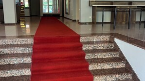 Een hotel lobby met een rode loper die leidt naar een receptiebalie, een trap en een groot beeld in een lijst aan de muur.