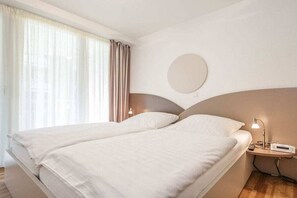 A hotel room with two beds, a bedside table with a lamp, a round mirror, and a window with curtains. - Holiday apartment with 2 bedrooms (Cuxhaven)