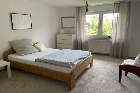 A bedroom with a wooden bed, a bedside table, a dresser, a window with curtains, and a framed picture on the wall.