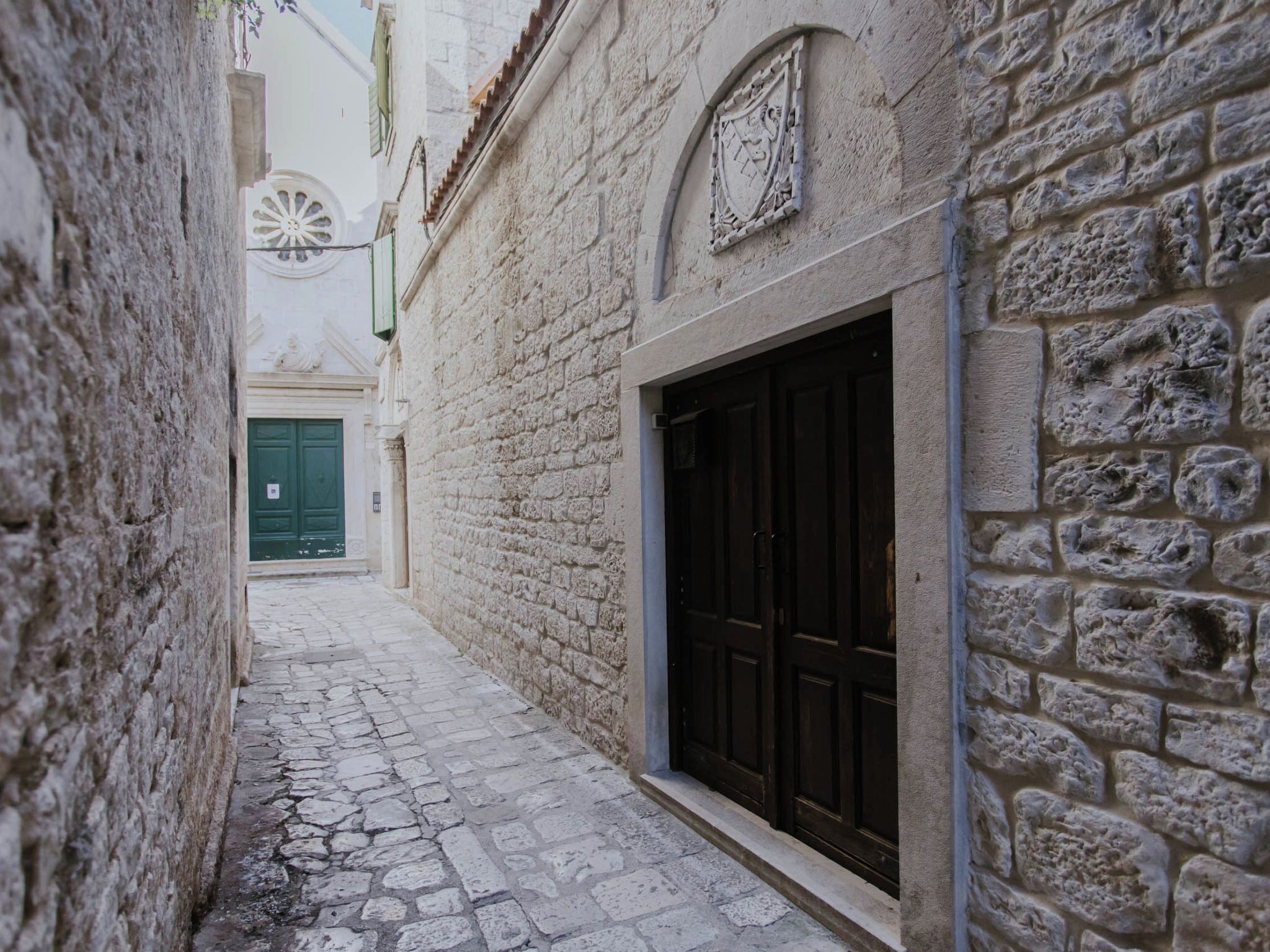 A narrow cobblestone street with stone buildings, a green door, and a coat of arms.