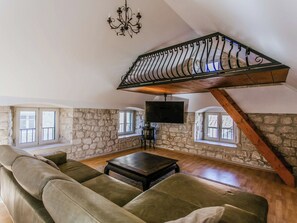 Ein Loft-Zimmer mit Steinsäulen, Holzbalkendecke, einer großen Couch, einem Couchtisch und einem an der Wand montierten Fernseher.