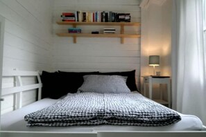 Ein kleines, ordentlich eingerichtetes Schlafzimmer mit einem Bett, einem Nachttisch mit Lampe und einem Regal mit Büchern.