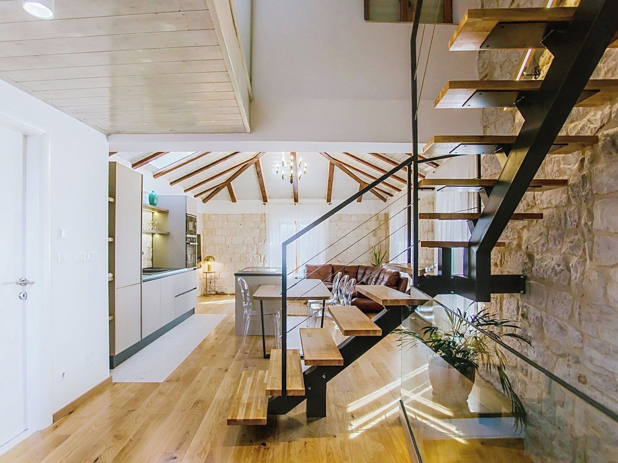 Eine moderne Küche mit weißen Schränken, ein Essbereich mit einem Holztisch und -stühlen sowie eine Treppe mit Glastrennwand.