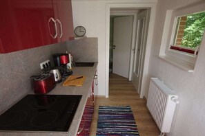 Une cuisine compacte équipée d’une cuisinière, d’un grille-pain et de placards rouges. Une fenêtre laisse entrer la lumière naturelle, et un radiateur est visible sur le mur.
