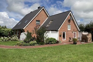 Une maison à deux étages, construite en briques, avec un toit en pente, un patio couvert et un petit jardin agrémenté de fleurs et d’arbustes.