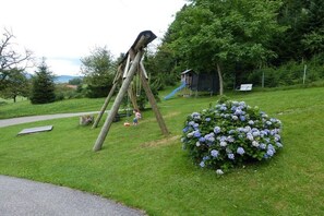 Un'area giochi con altalene e scivolo, immersa nel verde e con un cespuglio fiorito di fiori blu.
