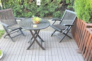 Eine Holzterrasse mit einem runden Tisch und zwei Stühlen, umgeben von einem Holzzaun und Grünpflanzen.